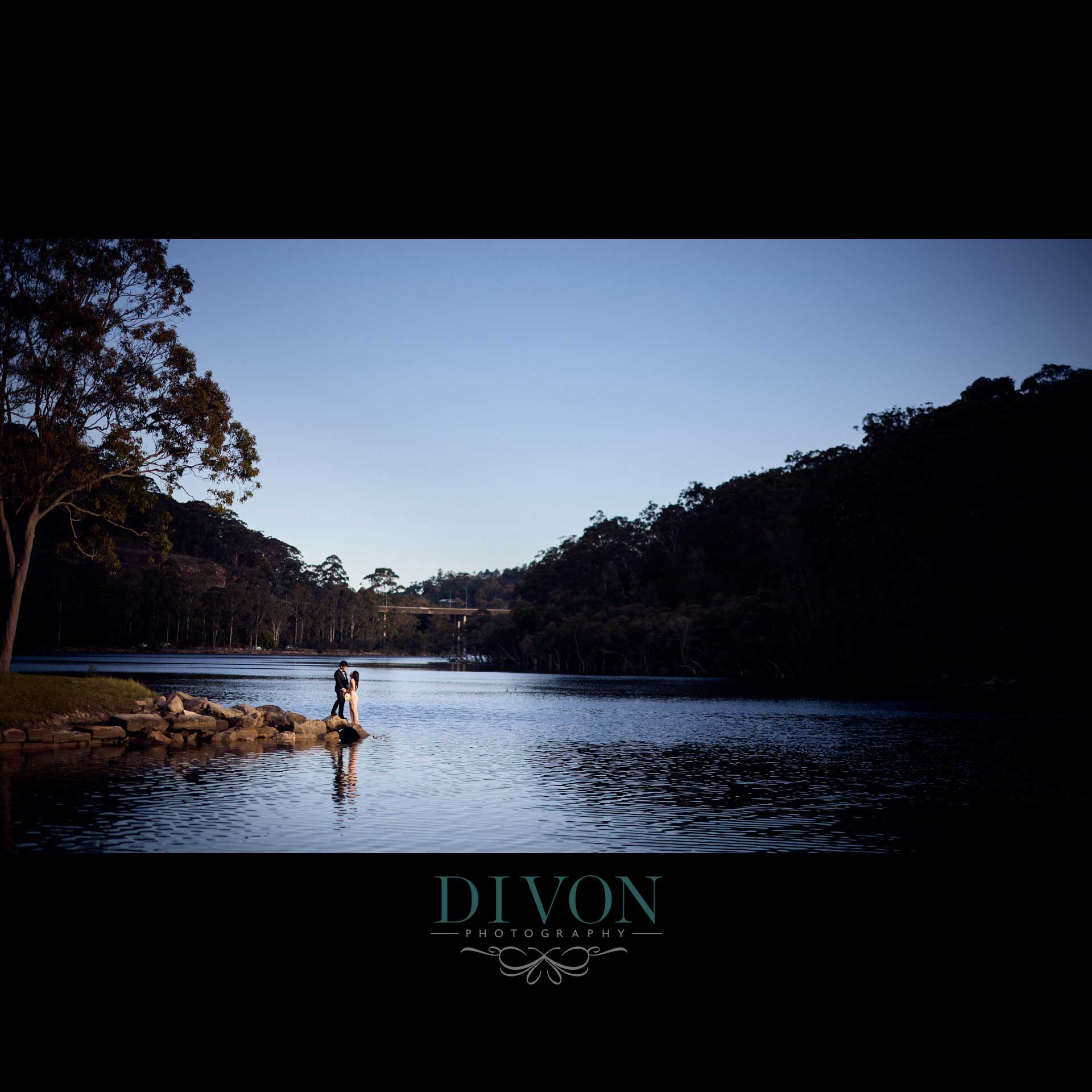 Ian and Romana standing together on rocks beside calm water at Davidson Park, framed by bushland and blue sky.